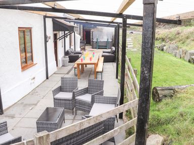 An outdoor patio area with a wooden dining table and benches cushioned chairs and sofas under a pergola at Maestyddyn Farm House in Clawddnewydd near Ruthin