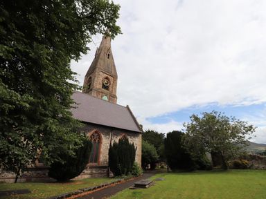 An old stone church with a tall steeple and clock surrounded by trees and grass at Maestyddyn Farm House in Clawddnewydd near Ruthin