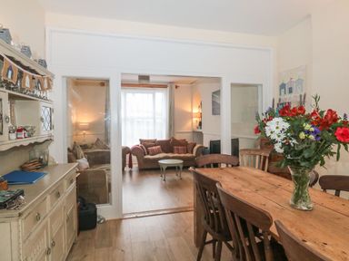 A dining room with a wooden table and chairs and a floral arrangement with a sitting room with a sofa and coffee table behind glass doors at Dunholme House in Teignmouth
