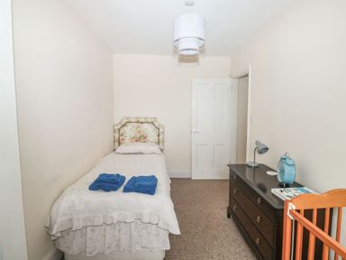 A bedroom with a single bed with floral headboard blue towels on bed a dark dresser with a lamp clock and books and a wooden crib at Dunholme House in Teignmouth