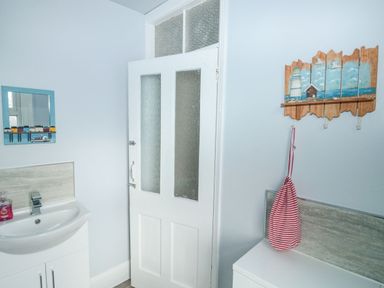 A bathroom with a white sink vanity with soap a white door with frosted glass panels a wooden wall hook with a lighthouse painting and a red and white striped bag hanging at Dunholme House in Teignmouth