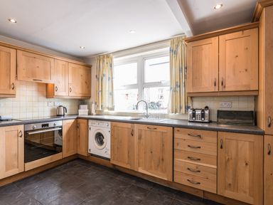 A kitchen with wooden cabinets a washing machine a toaster a kettle and a window with curtains at Fisherman's Cottage in Seahouses