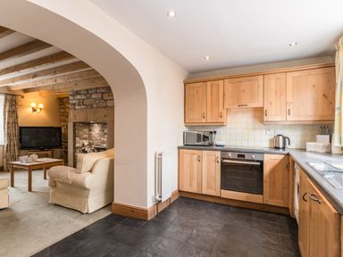 A kitchen with wooden cabinets and appliances next to a living room with armchairs and a TV at Fisherman's Cottage in Seahouses