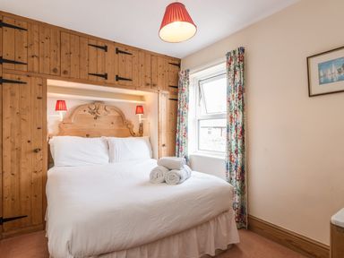 A bedroom with a bed white linens wooden headboard built in wooden cupboards window with floral curtains and red lampshades at Fisherman's Cottage in Seahouses