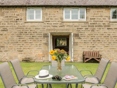 A garden table with chairs a vase of sunflowers a white hat binoculars and magazines outside a stone house with open doors at Brian's Cottage in Alnmouth