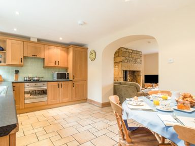 A kitchen with wooden cabinets and a dining table set for breakfast next to a living room with a stone fireplace at Brians Cottage in Alnmouth
