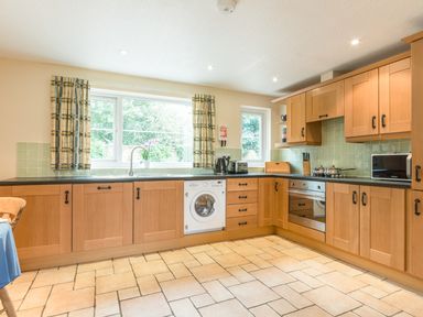 A kitchen with wooden cabinets a washing machine oven microwave and windows with curtains at Brians Cottage in Alnmouth
