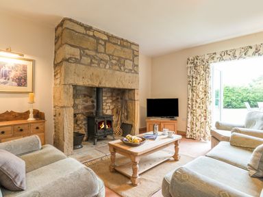A sitting room with a stone fireplace wooden furniture and floral curtains at Brian's Cottage in Alnmouth