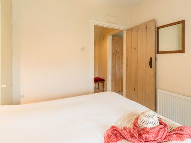 A bedroom with a bed, wooden door, mirror on the wall, radiator, and a hat with a red scarf on the bed at Brians Cottage in Alnmouth