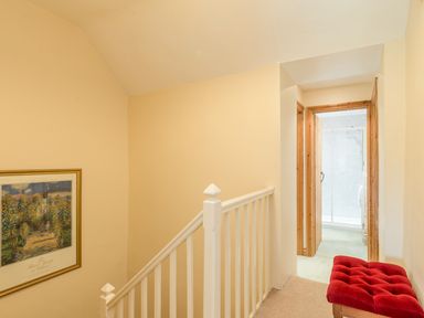 A hallway with a white railing a framed picture on the wall a wooden door leading to a bathroom and a red cushioned bench at Brian's Cottage in Alnmouth