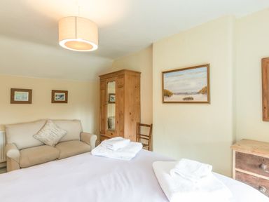 A bedroom with a bed, sofa, wooden wardrobe, chair, paintings on the walls, and folded towels on the bed at Brian's Cottage in Alnmouth