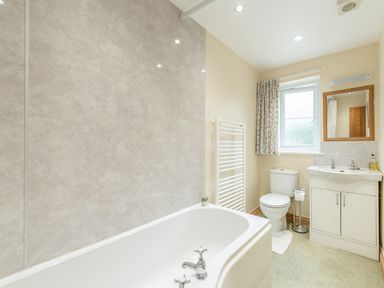A bathroom with a bathtub a toilet a sink with cabinet a window with a curtain and a towel radiator at Brian's Cottage in Alnmouth