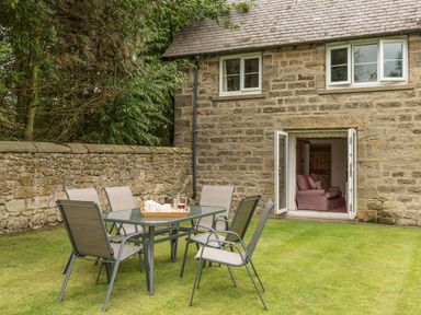 A patio table with six chairs on grass outside a stone house with open doors showing a sofa inside at Brians Cottage in Alnmouth