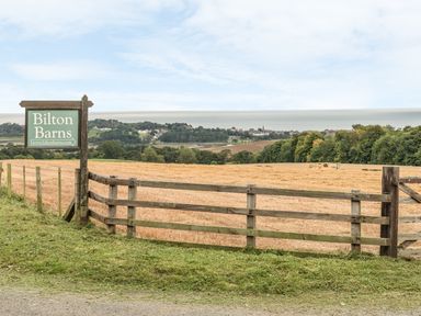 A wooden fence and a sign reading Bilton Barns beside a field with trees and buildings in the distance