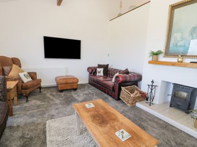 A living room with a brown leather sofa armchair and ottoman a wooden coffee table a flat screen tv mounted on the wall and a fireplace with a basket of firewood at Minerva in Trearddur Bay
