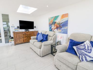 A living room with beige sofas with blue cushions a wooden TV cabinet and a colorful painting at Minerva in Trearddur Bay