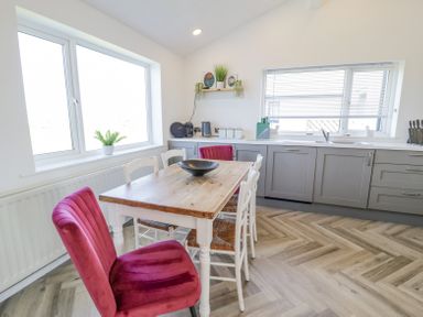 A kitchen with a wooden table surrounded by white and red chairs next to two windows and grey cabinets at Minerva in Trearddur Bay