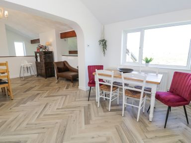 A dining area with a wooden table surrounded by white and red chairs next to a large window and a seating area with a brown chair and a wooden cabinet at Minerva in Trearddur Bay