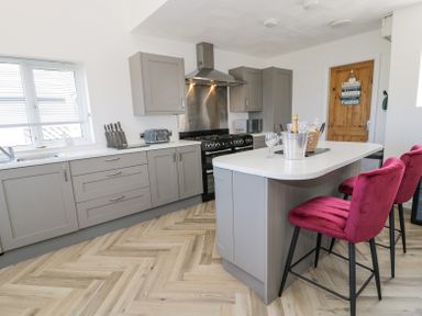 A kitchen with gray cabinets a stove and an island with two pink chairs at Minerva in Trearddur Bay