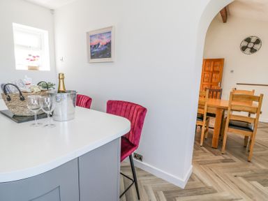 A kitchen island with two red chairs a bucket with a bottle and two glasses and a basket next to an archway leading to a dining area with a wooden table and chairs at Minerva in Trearddur Bay