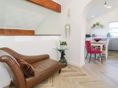 A brown leather chaise lounge with a brown pillow beside a glass side table with a plant in a room with wooden ceiling beams and a dining area with a table and chairs