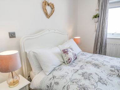 A bedroom with a white bed and floral bedding two bedside tables with lamps and a hanging plant at Minerva in Trearddur Bay