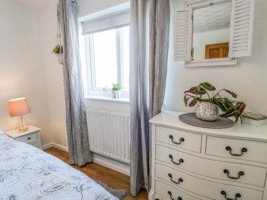 A bedroom with a white dresser a potted plant a window with curtains and a bedside table with a lamp at Minerva in Trearddur Bay