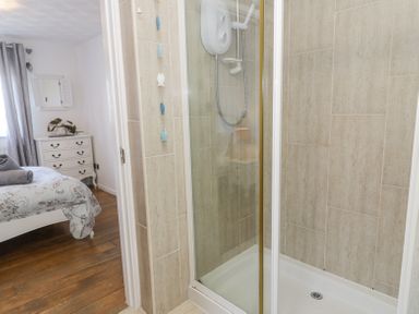 A shower area with a glass door and tiled walls next to a bedroom with wooden floors a white chest of drawers and a bed at Minerva in Trearddur Bay