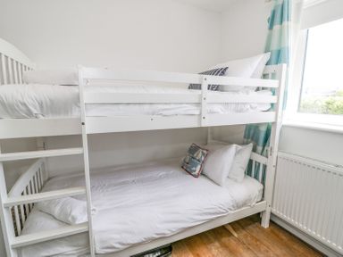 A white wooden bunk bed with white bedding and pillows in a room with a window and striped curtains at Minerva in Trearddur Bay
