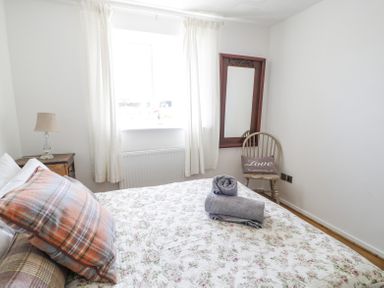 A bedroom with a bed covered in floral bedding pillows rolled towels a wooden chair with a love pillow a mirror and a window with white curtains at Minerva in Trearddur Bay
