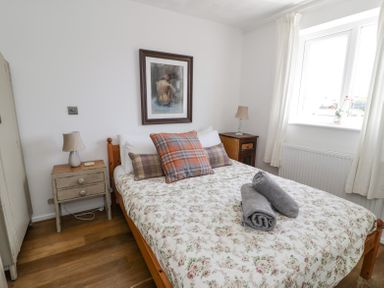 A bedroom with a wooden bed white floral bedspread plaid pillows bedside tables with lamps a framed painting and a window at Minerva in Trearddur Bay