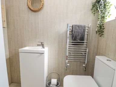 A small bathroom with a white sink unit a toilet a chrome towel rail with a grey towel and a basket with toilet paper at Minerva in Trearddur Bay