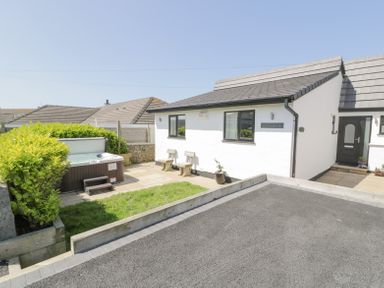 A white single-story house with a black door and two benches outside near a hot tub and garden at Minerva in Trearddur Bay