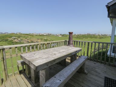 An outdoor wooden table with benches on a deck overlooking grassy fields and houses at Minerva in Trearddur Bay