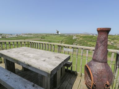 A wooden outdoor table with benches and a metal chiminea on a balcony overlooking a green landscape and houses at Minerva in Trearddur Bay