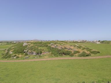 A grassy hill with shrubs and a dirt path in a rural area with houses in the distance at Minerva in Trearddur Bay