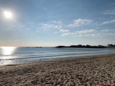 A sandy beach with calm sea water and small waves under a clear sky with scattered clouds at Minerva in Trearddur Bay