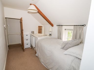 A small bedroom with a single bed beside a window with curtains a white dresser with a lamp and a mirror and a wooden door at Manor Farm Cottage in Swaffham