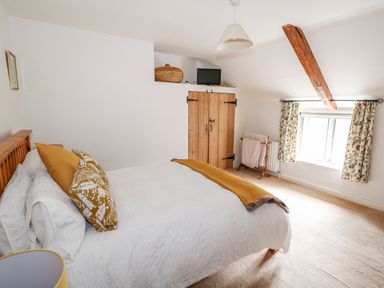 A bedroom with a bed with white bedding and yellow pillows a wooden wardrobe a TV a window with patterned curtains and a wooden beam on the ceiling at Manor Farm Cottage in Swaffham