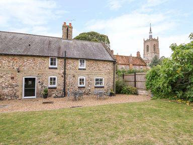 A stone cottage with a gravel patio and metal tables and chairs surrounded by grass and greenery at Manor Farm Cottage in Swaffham
