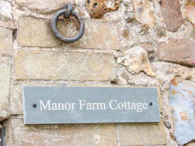 A stone wall with a metal ring above a sign that reads Manor Farm Cottage in Swaffham