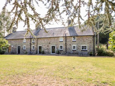 A stone cottage with multiple doors and windows seen from a grass lawn framed by tree branches at Manor Farm Cottage in Swaffham