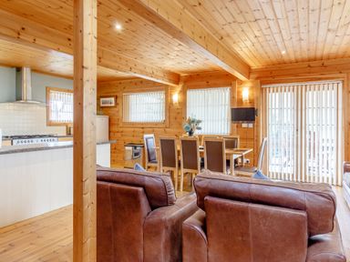 A wooden cabin interior with a dining table and chairs a kitchen area with appliances and two brown leather chairs at Jamaal Lodge in South Broomhill near Amble