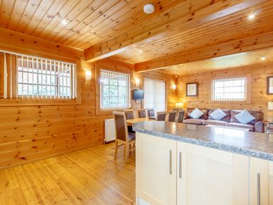 An open living space with a wooden dining table and chairs a leather sofa with blue cushions a countertop and wood paneled walls and ceiling at Jamaal Lodge in South Broomhill near Amble