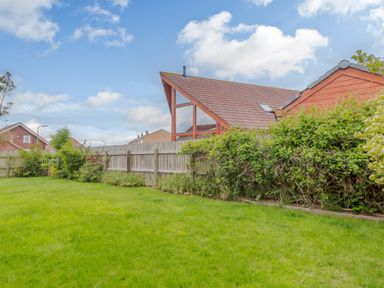 A backyard with green grass wooden fence and bushes near a house with large triangular windows at Jamaal Lodge in South Broomhill near Amble