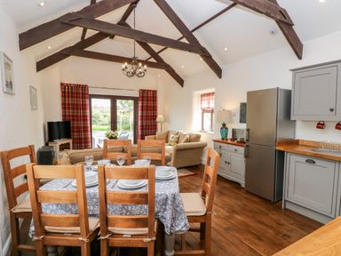 A dining table with six chairs set with plates and glasses in an open plan kitchen and living area with wooden beams on the ceiling at Potters Barn in Begelly near Kilgetty