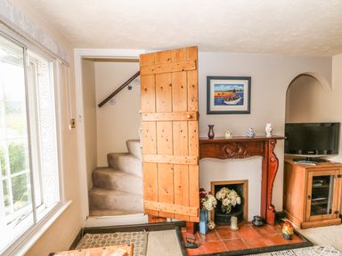 A room with a wooden door open to carpeted stairs a fireplace with decorative items a television on a wooden cabinet and a window with a view outside at Megs Cottage Norwich