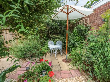 A small garden patio area with two white metal chairs a table and a large white umbrella surrounded by plants and flowers at Megs Cottage in Norwich