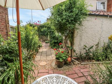 A garden area with plants flowers a white metal table an umbrella and a paved walkway at Megs Cottage in Norwich