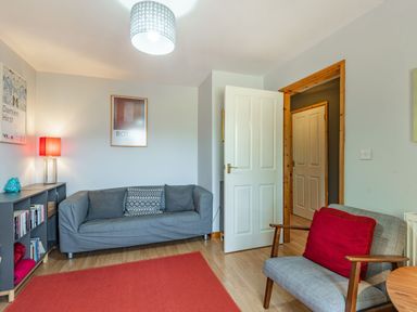 A sitting room with a grey sofa, an armchair with a red cushion, a red rug, a bookshelf, and a standing lamp at Gorwel in Llan Ffestiniog near Blaenau Ffestiniog